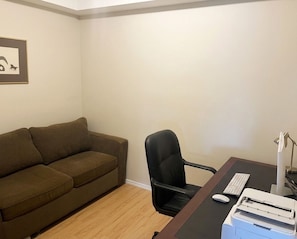 Een kamer met een bruine bank, een zwarte bureaustoel, een bureau met een computer, een printer en een lijst met een afbeelding aan de muur.