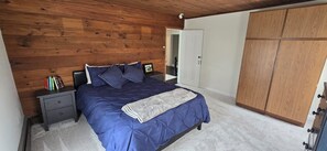 Un dormitorio con techo de madera, una cama con cubrecama y almohadas azules, una mesita de noche con libros y un armario.
