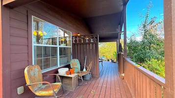 A wooden deck with outdoor furniture, including chairs and a table, under a covered area with a view of greenery.