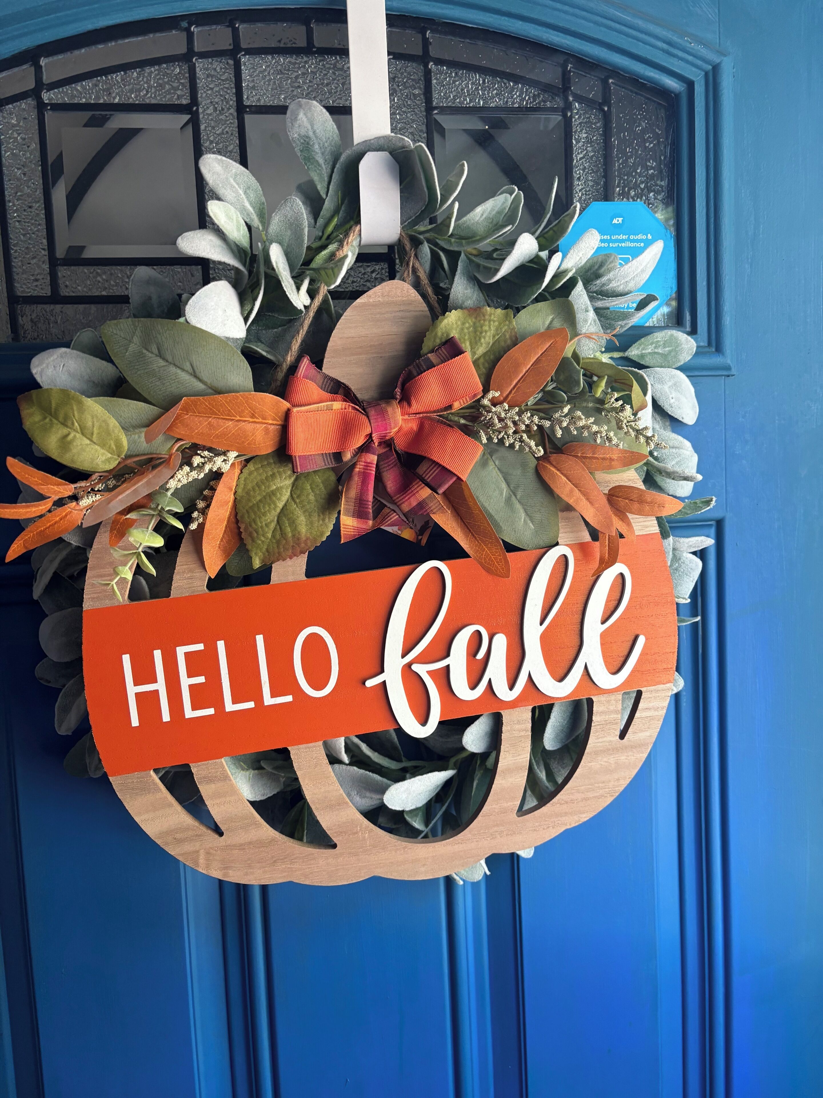 Ein hölzerner Kranz mit einem Schild, auf dem 'HELLO fall' steht, hängt an einer blauen Tür.