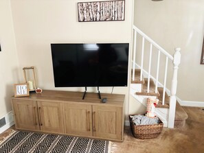 A living room with a flat-screen TV on a wooden cabinet, a framed picture above the TV, a staircase with a white railing, and a woven basket on the floor. - Cozy Comfy Pet Friendly House in a Quiet Neighborhood  (Wilmington)