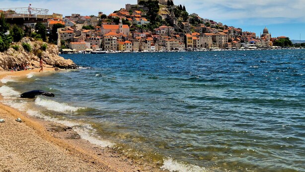 Una cittadina costiera con edifici su una collina, una spiaggia e un mare tranquillo.