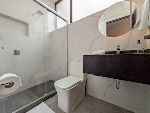 A modern bathroom with a glass shower enclosure, a toilet, a sink with a dark countertop, and a round mirror. - Casa Heredia 1Bed Studio (Guadalajara)