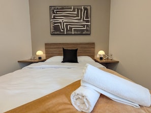 A hotel room with a bed, two bedside tables with lamps, a black pillow, a white towel, and a wall art piece. - Casa Heredia 1Bed Studio (Guadalajara)