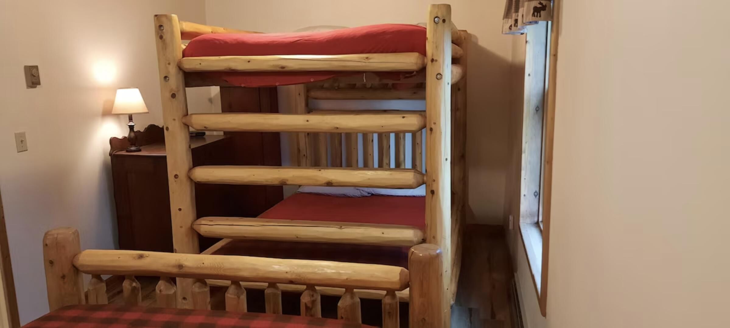 Un lit superposé en bois avec un matelas rouge, une table de chevet avec une lampe et une petite fenêtre.