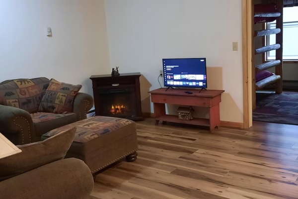 Natural light brightens the living room’s wide plank wood floor, with a small TV and electric fireplace in view.