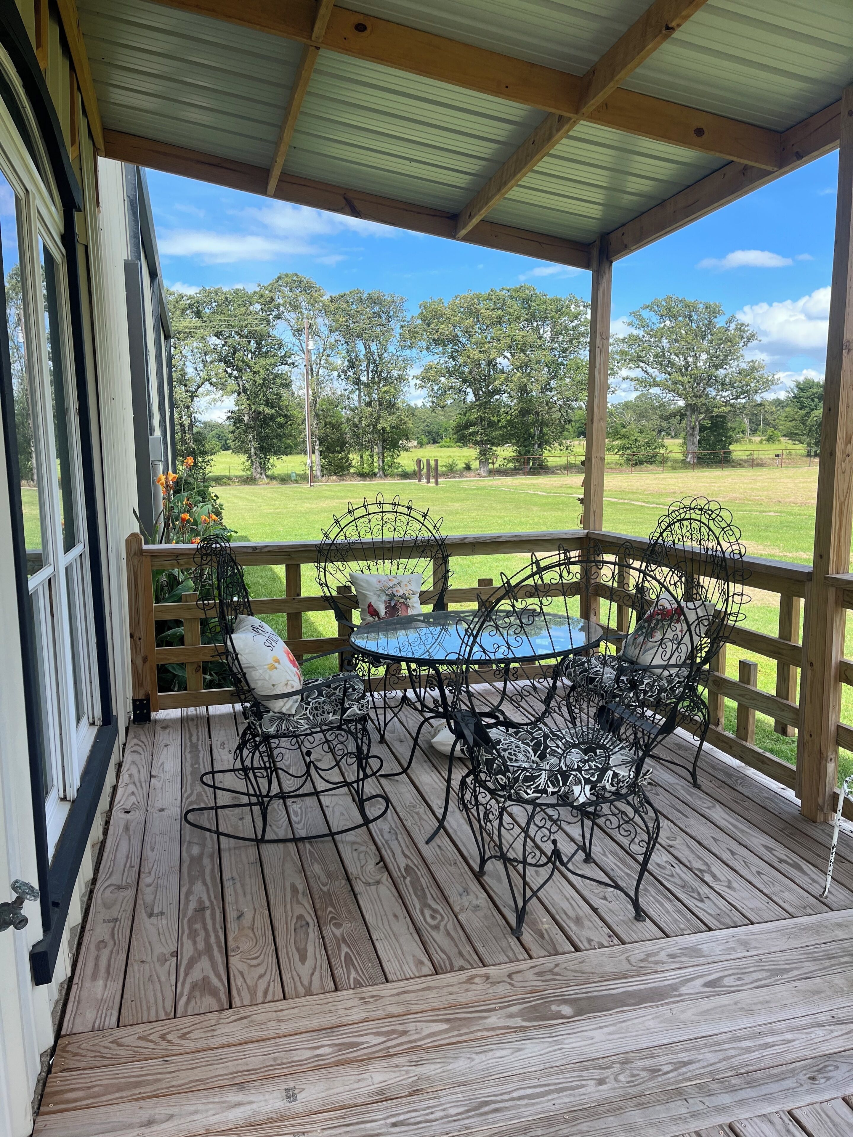 A covered porch with a wooden table and chairs, a view of a grassy field, and a wooden fence.