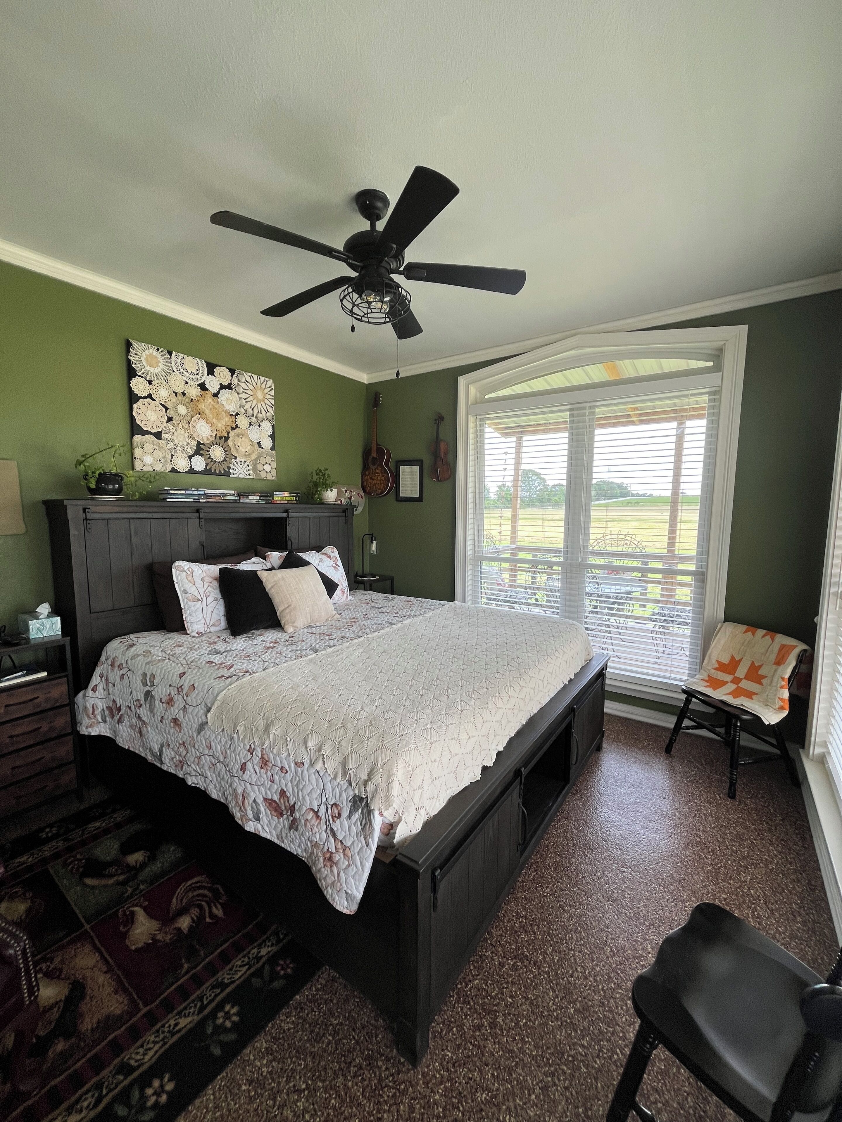 A bedroom with a bed, a ceiling fan, a window with blinds, and a view of a green landscape.