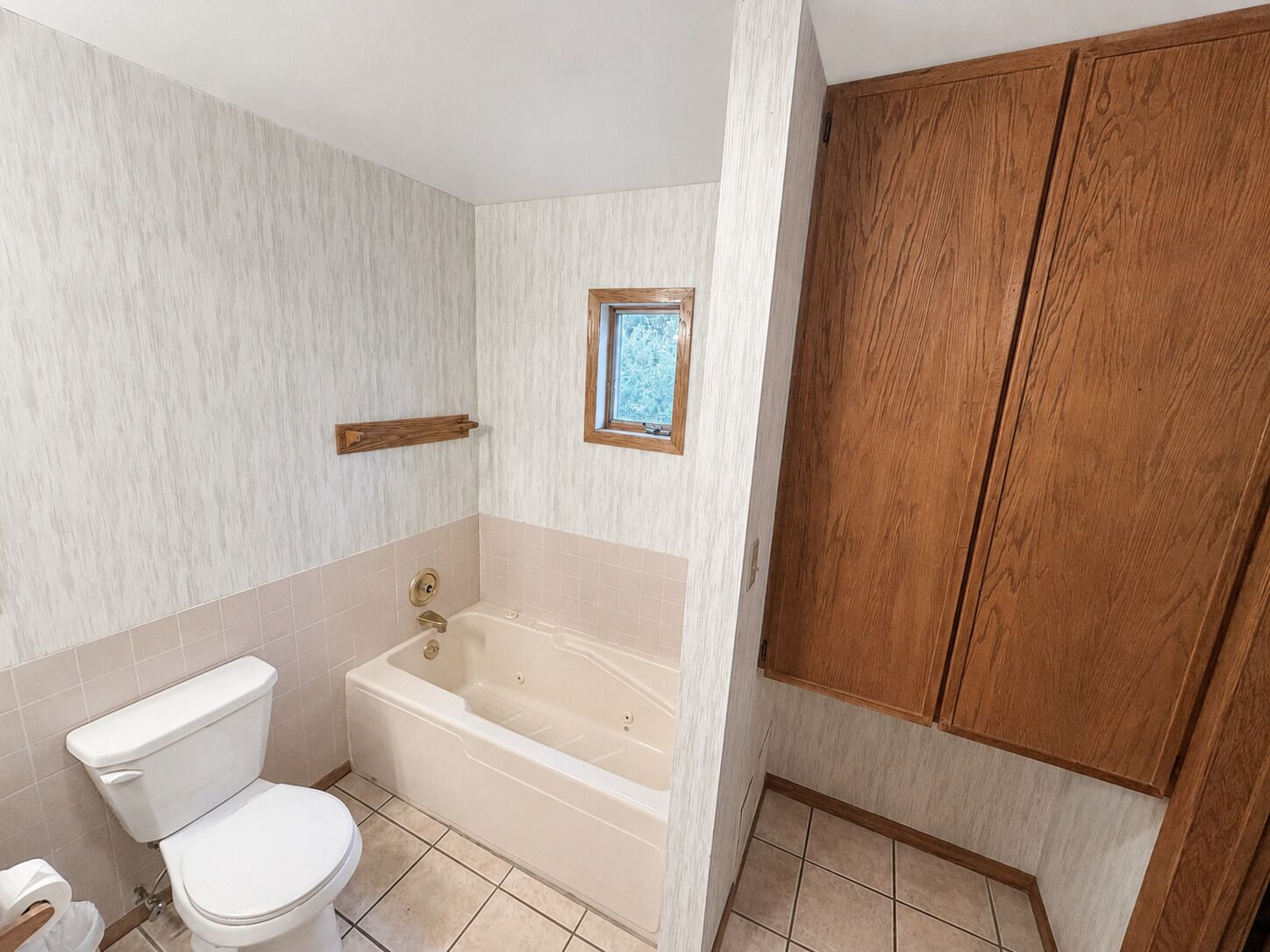 A bathroom with a toilet, bathtub, wooden cabinets, and a window.