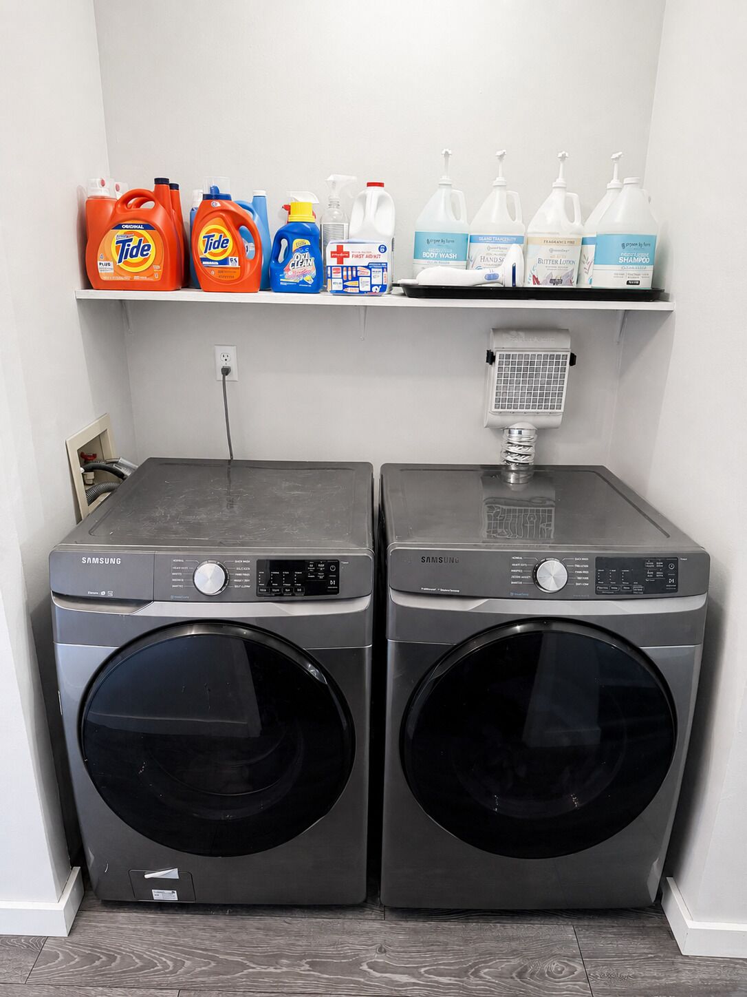 Een wasruimte met twee Samsung wasmachines, een plank vol schoonmaakmiddelen en een boiler.