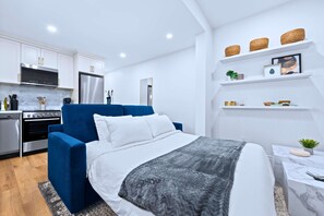 Une chambre moderne avec un lit, un fauteuil bleu, un petit bureau et un coin cuisine doté de meubles blancs et d’appareils électroménagers.