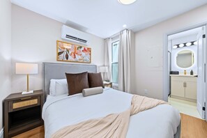 Une chambre d’hôtel comprenant un lit, une table de chevet, une lampe, un climatiseur, un tableau, une fenêtre avec des rideaux et une salle de bain avec un miroir et des luminaires.