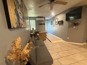 A living room with a gray sofa, a ceiling fan, a flat-screen TV, and a dining area with a table and chairs.