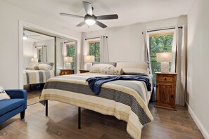 Quarto com cama, ventoinha no teto, mesa de cabeceira, cômoda e janela com cortinas.