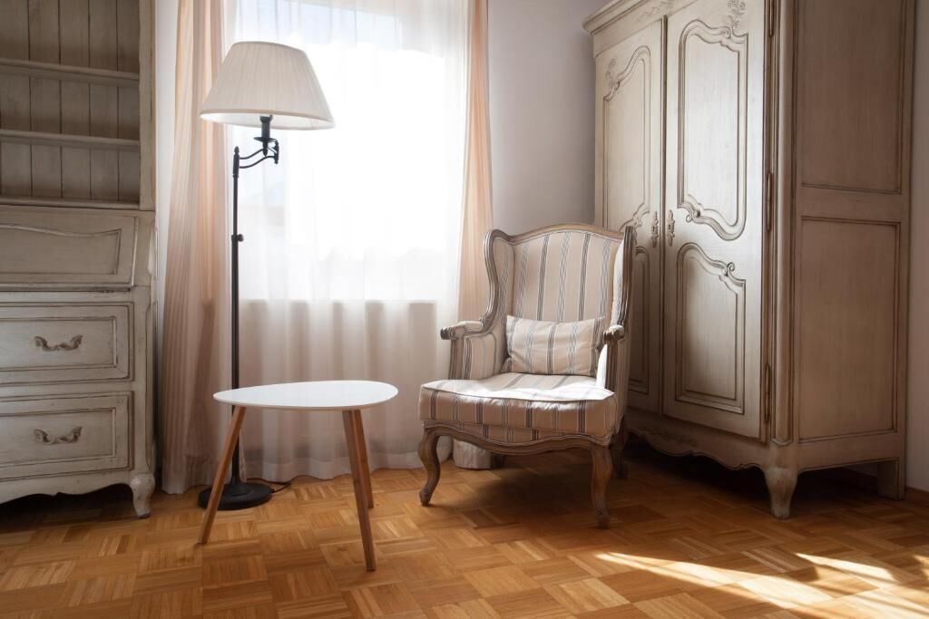 Una habitación con suelo de madera, una mesa blanca, un sillón a rayas, una lámpara y un armario blanco.
