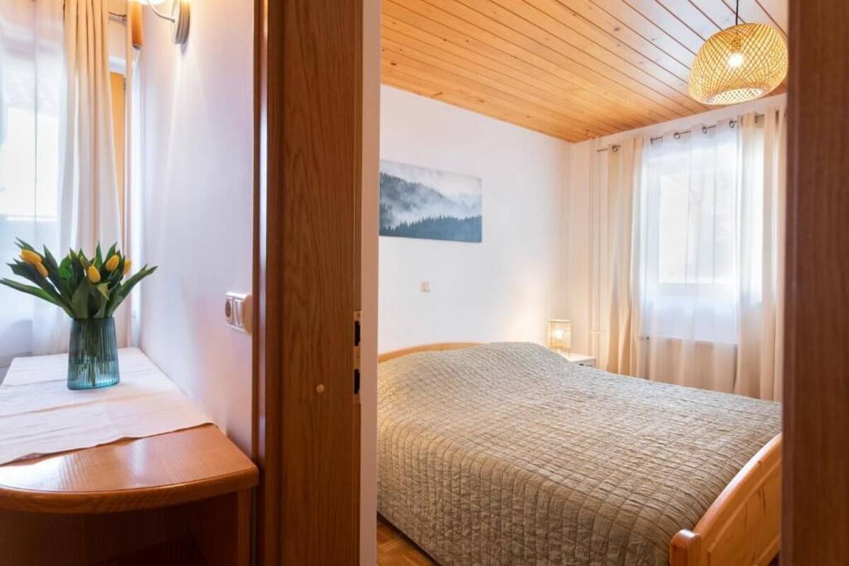 A bedroom with a bed, a bedside table, a window with curtains, a painting on the wall, and a wooden ceiling.