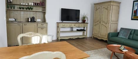 A living room with a wooden dining table, a green sofa, a television, and a cabinet with kitchenware.