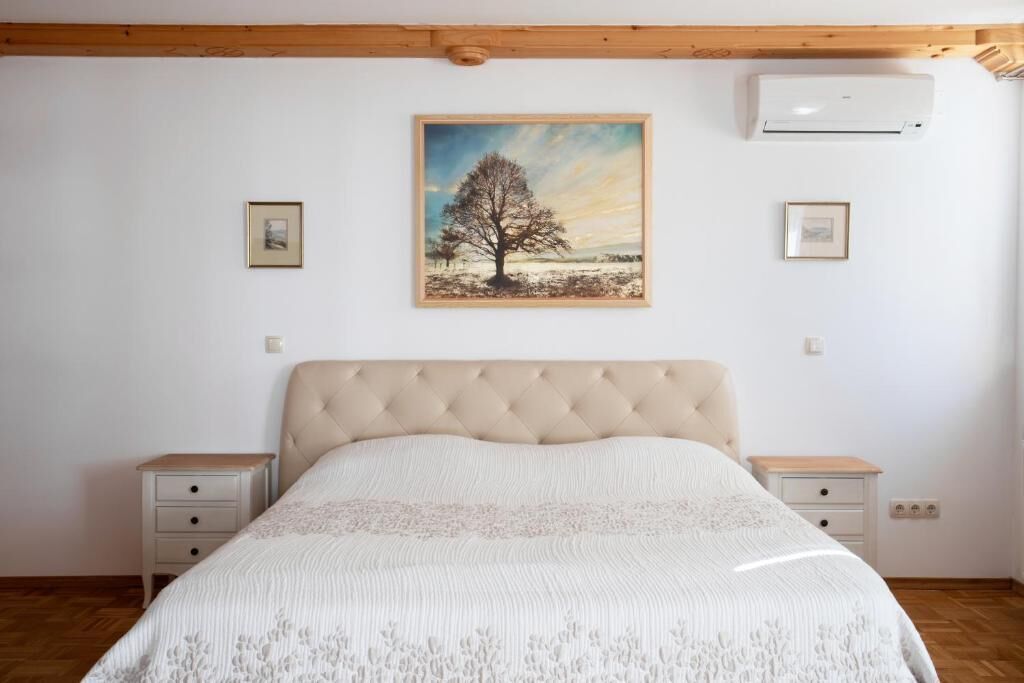 A bedroom with a tufted headboard, a large bed with a white textured bedspread, two bedside tables with drawers, and a framed landscape painting on the wall.