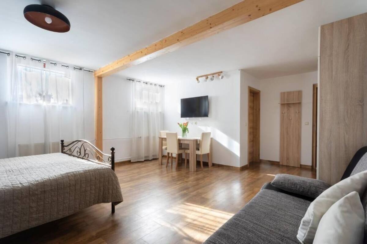 A room with a bed, a sofa, a TV, a desk with chairs, and wooden beams on the ceiling.