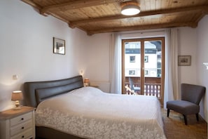 Un dormitorio con una cama, una silla, una mesita de noche y un ventanal con vistas a edificios.