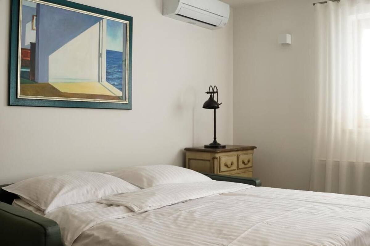 A bedroom with a bed, a nightstand, a lamp, a painting, and an air conditioning unit.
