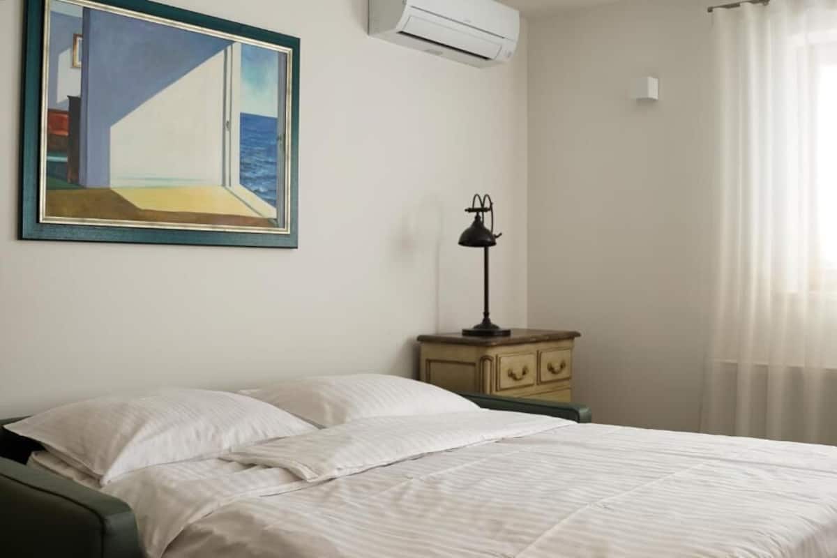A bedroom with a bed, a nightstand, a lamp, a painting, and an air conditioning unit.