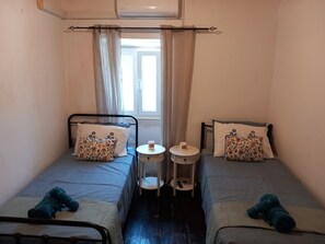 Una habitación pequeña y estrecha con dos camas individuales, cada una con una almohada y una mesita de noche.