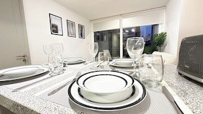 Mesa posta com pratos, copos e talheres, com vista para a paisagem urbana pela janela.