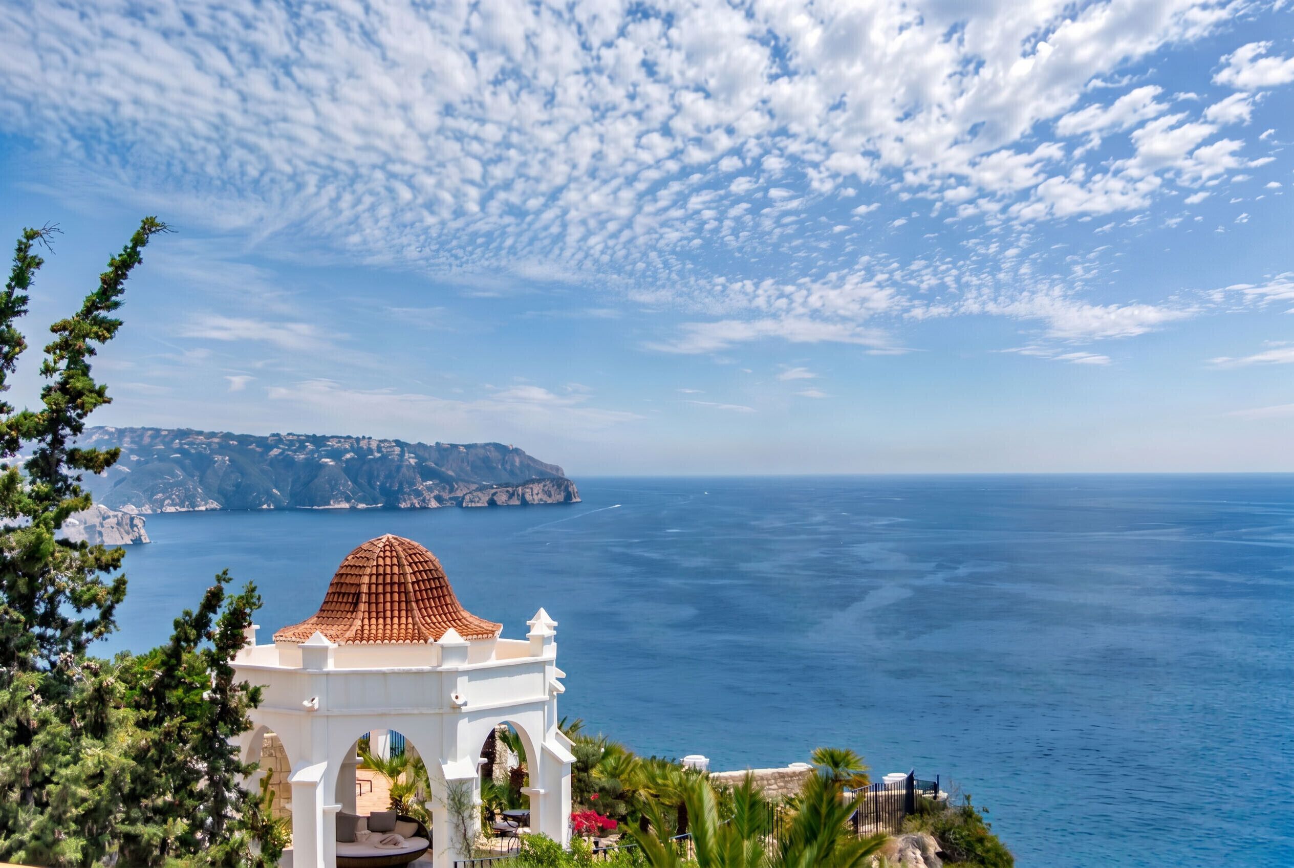 A coastal view with a white building featuring a red-tiled dome, overlooking the sea and a distant island.