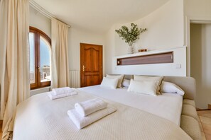 Ein großes Bett mit weißer Bettwäsche und Handtüchern, eine Holztür, ein an der Wand befestigtes Regal mit Dekoration und ein Fenster mit Vorhängen.