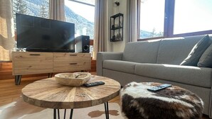 A living room with a sofa, a round wooden coffee table, a television on a wooden cabinet, and a view of mountains through large windows. - Pragelato Weekend (pragelato)