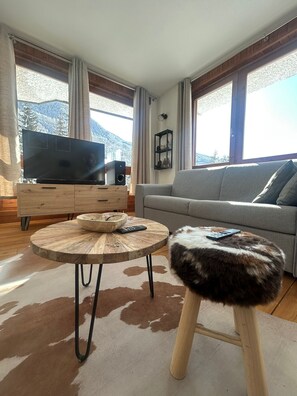 A living room with a sofa, a round wooden coffee table, a television on a wooden cabinet, and a view of mountains through large windows. - Pragelato Weekend (pragelato)