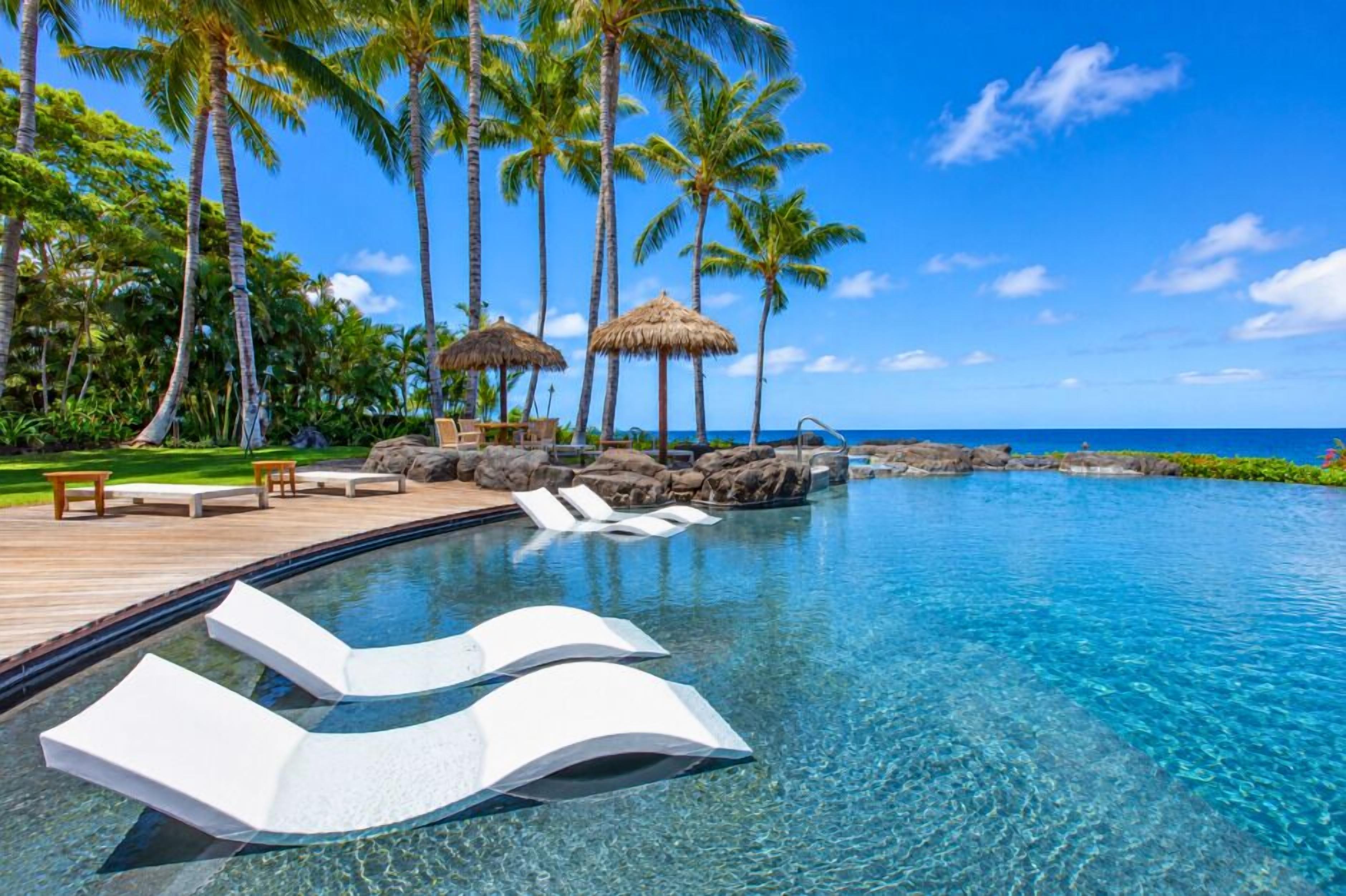 A clear blue pool with lounge chairs, palm trees, and a wooden deck.