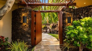 A stone pathway leading to a wooden gate with a tropical garden on either side. - One Ocean Estates by KBM (Waikoloa)
