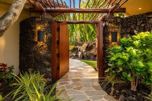 A stone pathway leading to a wooden gate with a tropical garden on either side. - One Ocean Estates by KBM (Waikoloa)