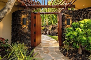 A stone pathway leading to a wooden gate with a tropical garden on either side.