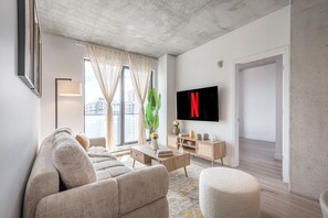 Un soggiorno moderno con divano, tavolino da caffè e poggiapiedi, una TV a parete e un'ampia finestra con tende.