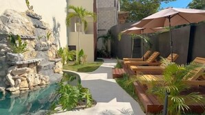 Un espace piscine doté de chaises longues, d’une cascade artificielle en roches et d’un chemin menant à un bâtiment.