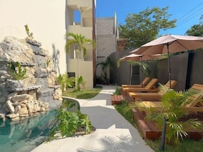A pool area with lounge chairs, a rock waterfall feature, and a pathway leading to a building. - Modern Swim-Up Studio by the Pool - Mizu 20 (Tulum)