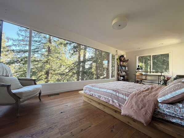 A bedroom with a large window, wooden flooring, a bed with patterned bedding, a chair, a desk, and a bookshelf.