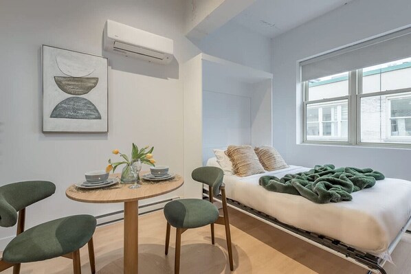 Ein modernes Schlafzimmer mit Bett, Fenster, Esstisch mit Stühlen und einer Wand-Klimaanlage.