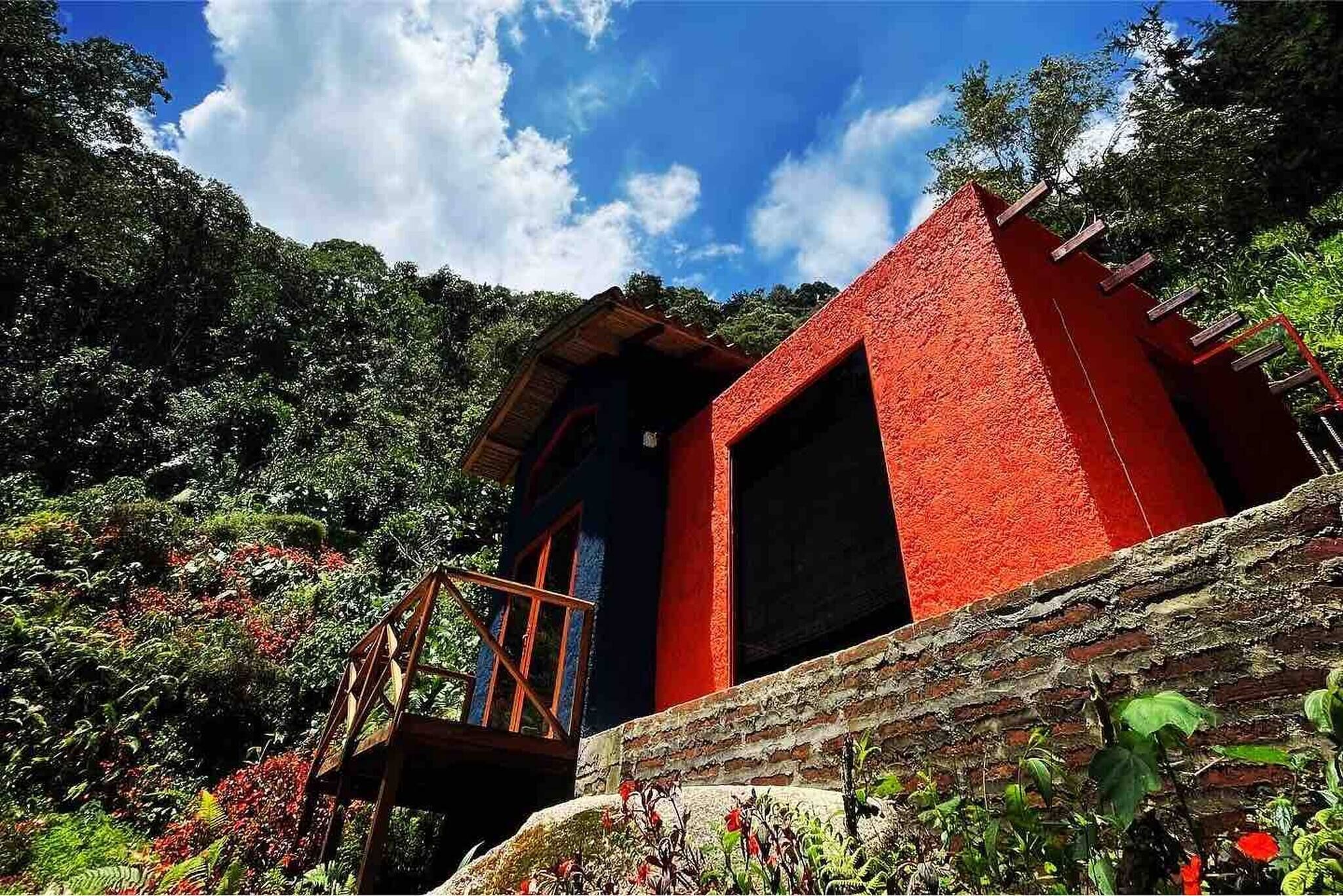 Un edificio rojo con una escalera de madera que lleva a una puerta, rodeado de abundante vegetación.