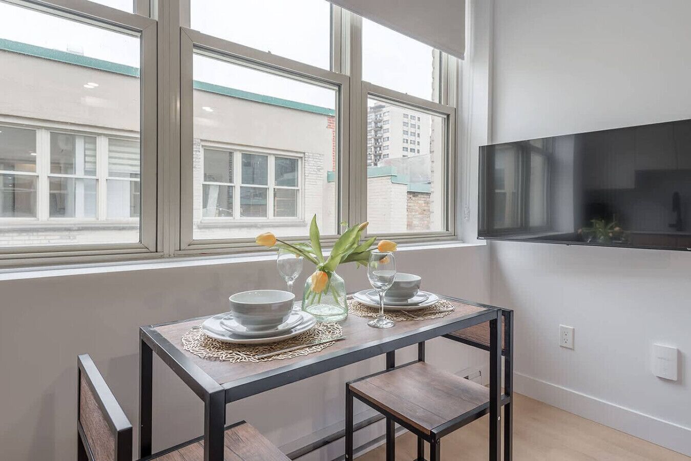 Un coin repas avec une table dressée pour deux personnes, un vase rempli de fleurs et un téléviseur fixé au mur.