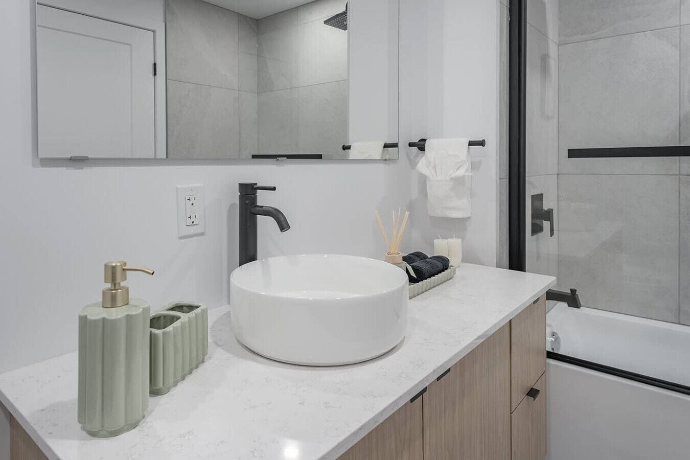 Une salle de bain moderne avec un grand lavabo blanc, un robinet noir et un miroir au-dessus. On y trouve deux distributeurs verts et une bougie sur le comptoir.