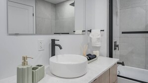 Une salle de bain moderne avec un grand lavabo blanc, un robinet noir et un miroir au-dessus. On y trouve deux distributeurs verts et une bougie sur le comptoir.