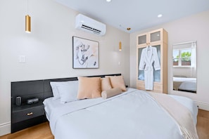 Une chambre moderne comprenant un lit, une table de chevet, un appareil de climatisation, des œuvres d’art et une robe suspendue.