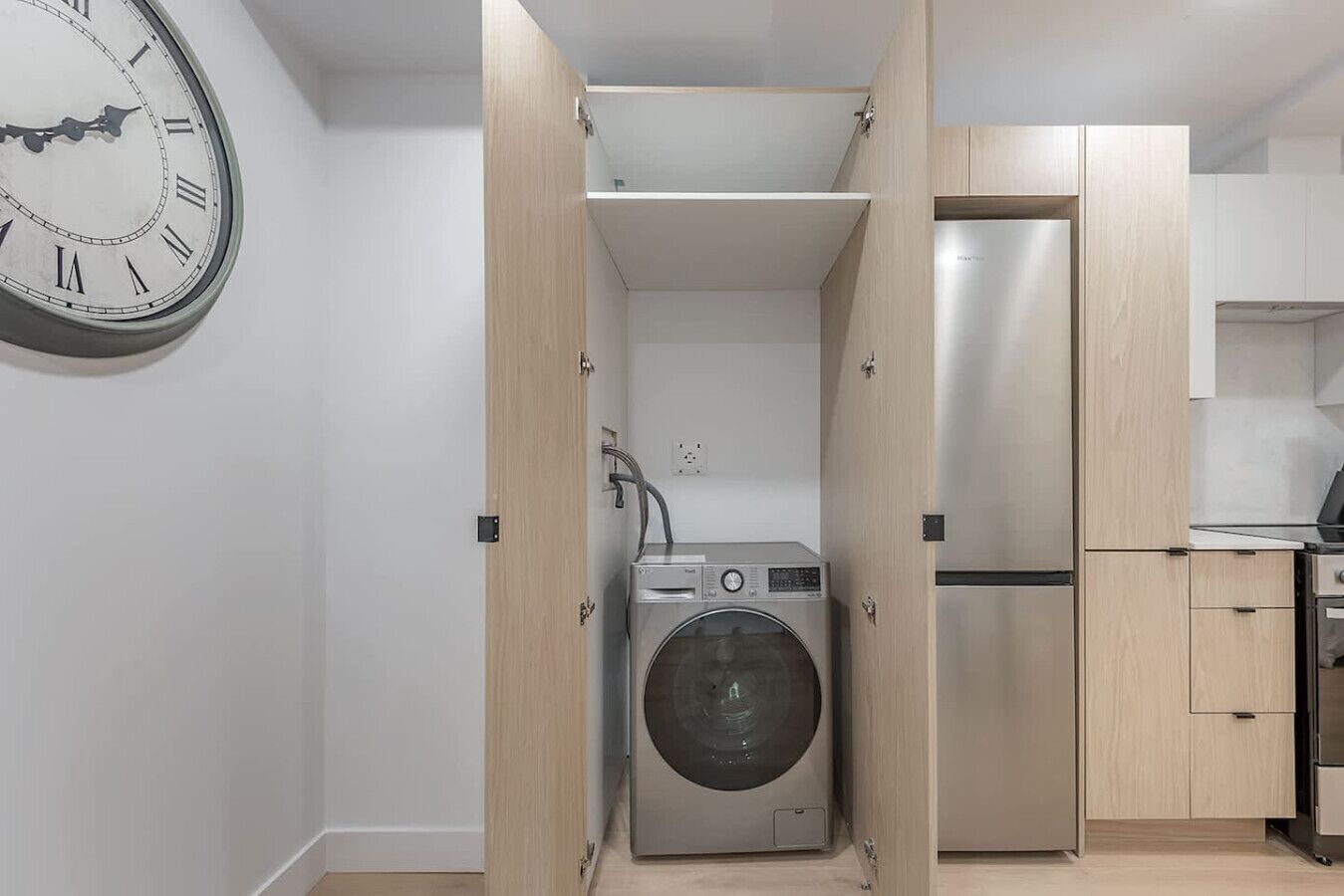 Une cuisine compacte équipée d’un lave-linge et d’un sèche-linge intégrés, d’un réfrigérateur en acier inoxydable et d’une horloge murale.