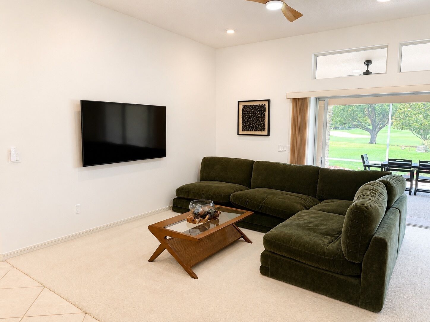 A living room with a flat-screen TV, a green sofa, a glass coffee table, and a ceiling fan.