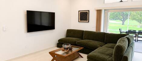 A living room with a flat-screen TV, a green sofa, a glass coffee table, and a ceiling fan.