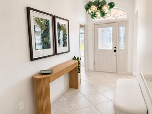 A hallway with a wooden console table, two framed artworks, a potted plant, and a white door with a stained glass window. - Golf Course Retreat in The Villages with Water Views, Lanai & Prime Location (Lady Lake)